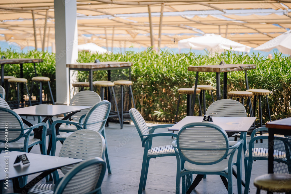 Obraz premium Outdoor terrace with modern tables, chairs, and bar stools under large sunshades. A stylish empty seating area in a sunny resort or beachside café.