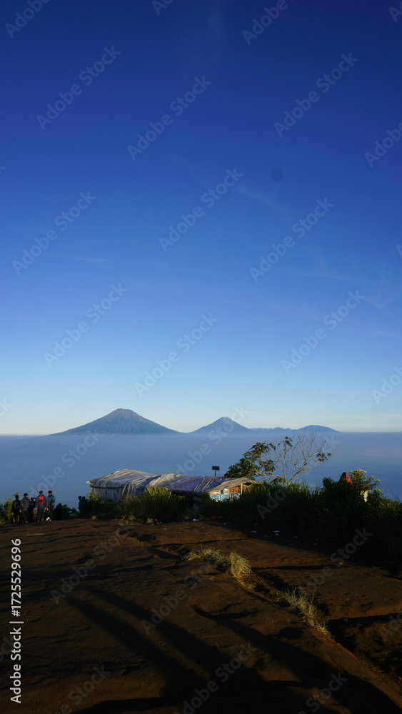 Obraz premium Panoramic view of a mountain landscape at sunrise, with a sea of clouds filling the valley below and a clear blue sky above.
