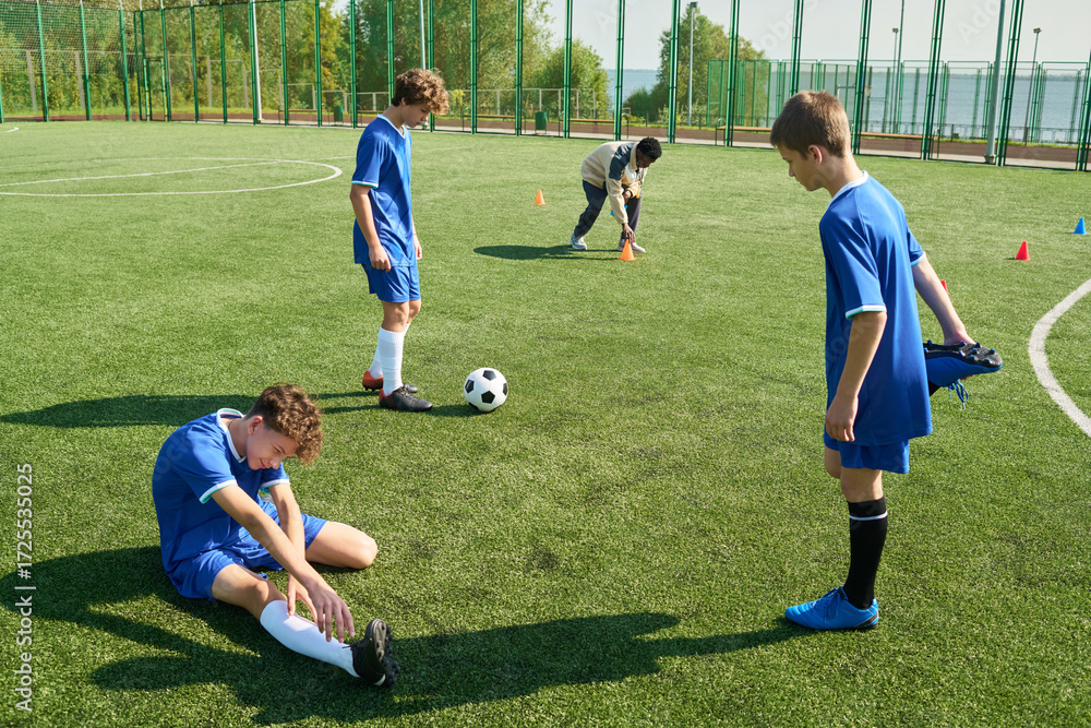 Obraz premium Group of teenage boys playing soccer on outdoor field, one teenager sitting on grass stretching leg while others standing nearby, soccer ball and cones visible in background