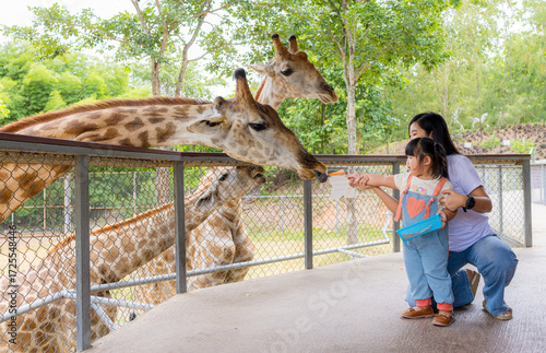 Canvas Print Asian mother and little daughter feed carrots to giraffes at a zoo during a summer holiday,Happy family, young mother with one child, cute toddler feeding a giraffe during a visit to the city zoo