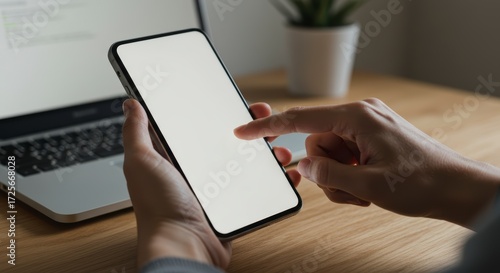 Person using a smartphone with a blank white screen mockup at a wooden desk with a laptop.