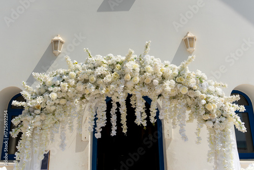 A stunning floral archway adorned with white roses and cascading flowers stands at the entrance of a bright venue during a wedding ceremony. The elegant decorations create a romantic atmosphere.