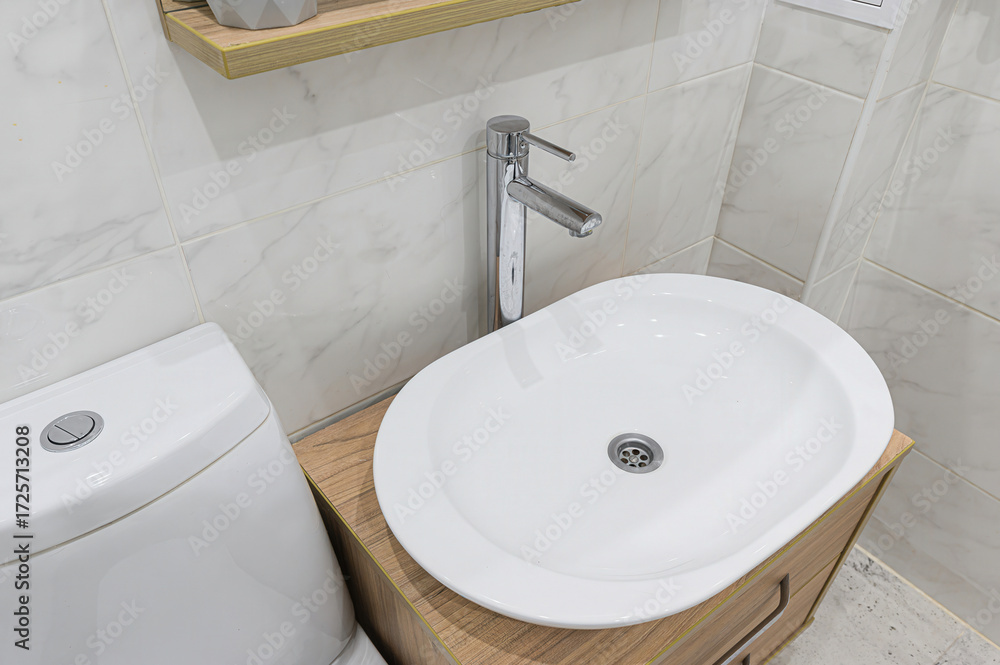 Fototapeta premium A close-up shows a modern bathroom setup with a white oval sink, chrome faucet, toilet, and tiled walls