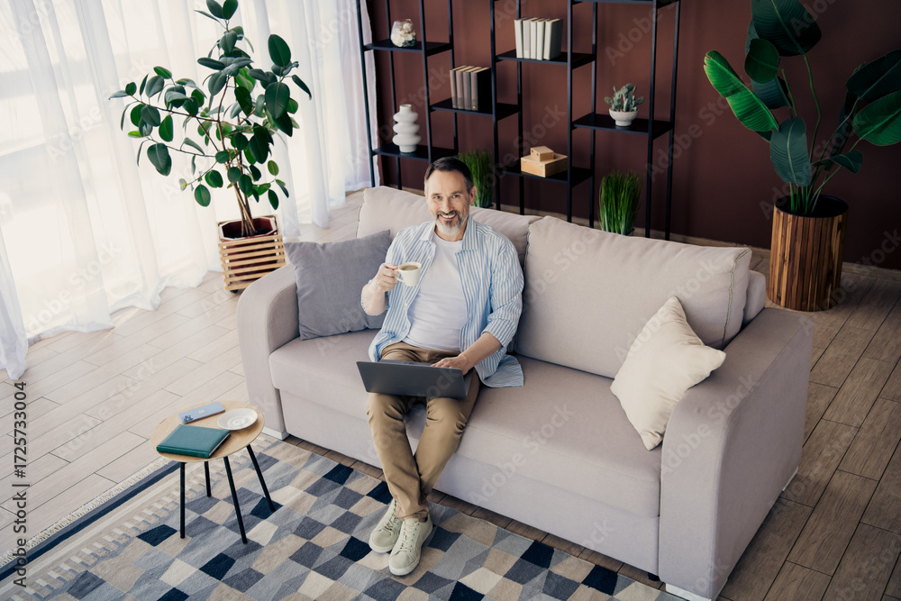 Obraz premium Confident mature man working on laptop while enjoying coffee in a modern and stylish home interior