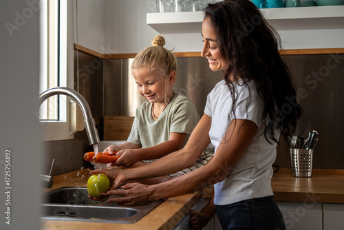 Φωτογραφία Young boy washing fresh carrot under running water with his mother holding green