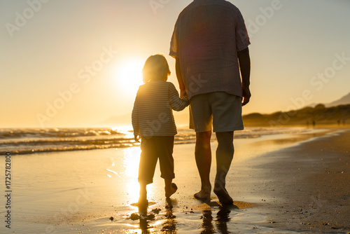 Tableau sur toile Back view of grandfather and grandson walking barefoot along the shoreline at sunset, hand in hand, enjoying a peaceful evening together on a quiet beach