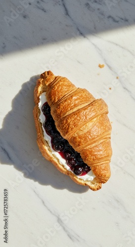 A croissant filled with white cream and dark berries sitting on a marble surface in sunlight
