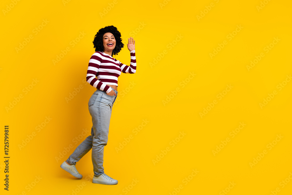 Fototapeta premium Cheerful young woman with curly hair in casual style waving on yellow background