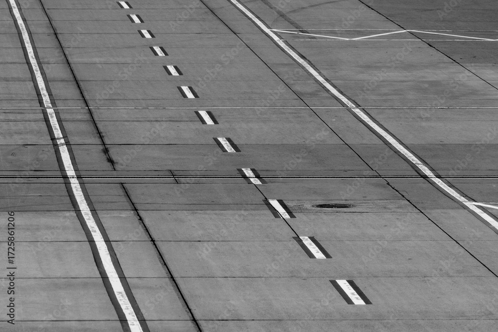 Fototapeta premium A black-and-white view of an airport tarmac road featuring parallel white line markings, emphasizing operational layout under overcast skies. Stock photo