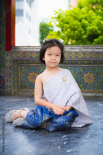 Beautiful traditional portrait of young Asian child girl wearing an Thai traditional dress Sabai and loincloth with silver sparkling shoes and brooch. Kid is sitting politely on patterned stone floor.