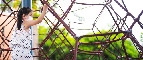 Close up portrait of cute Asian child wearing protective white surgical face mask. Girl are climbing on a rope net in an outdoor playground. health, safety, and childhood during the flu pandemic.