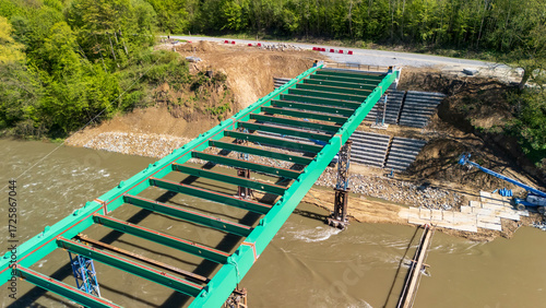 Aerial view of a partially constructed green steel bridge over a river, surrounded by lush greenery. Heavy machinery is visible nearby, indicating ongoing construction and development.