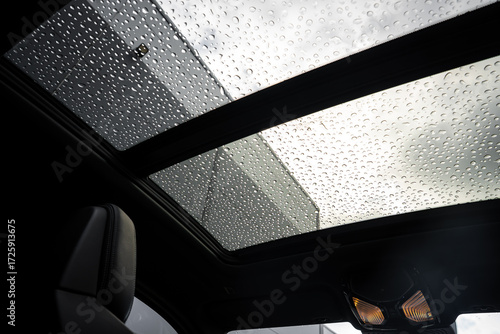 Foto Interior view of a BMW X4 focusing on its panoramic sunroof covered in rain droplets, with a cloudy sky and modern building visible in the background
