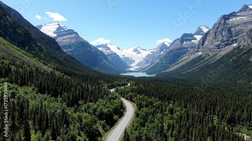 Aerial view of a scenic road winding through a lush green forest in glacier national park, montana, with snowcapped mountains in the background on a sunny day