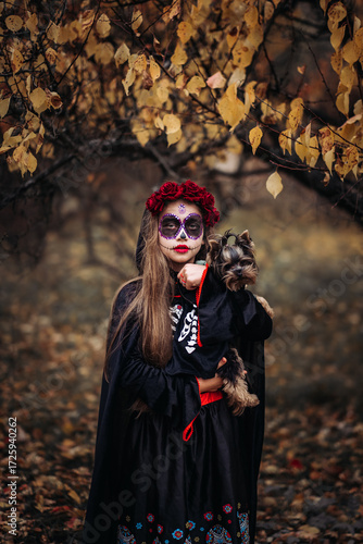 A young woman in a Day of the Dead costume: purple and violet skull makeup, red roses on a wreath, and a black cloak. She held a small dog against the backdrop of an autumn park with fallen leaves.