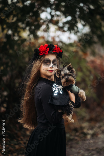 A young woman in a Day of the Dead costume: purple and violet skull makeup, red roses on a wreath, and a black cloak. She held a small dog against the backdrop of an autumn park with fallen leaves.