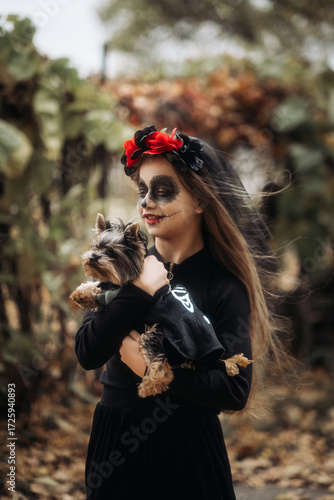 A young woman in a Day of the Dead costume: purple and violet skull makeup, red roses on a wreath, and a black cloak. She held a small dog against the backdrop of an autumn park with fallen leaves.