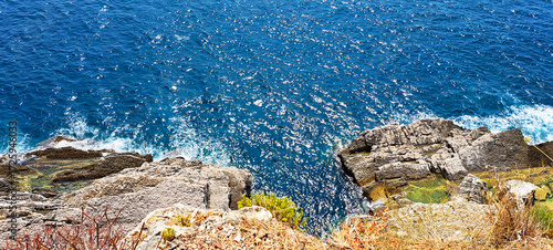 Turquoise seawater laps against rugged layered rock formations on a steep coastline, showcasing striking geological textures and the dynamic meeting of land and sea.