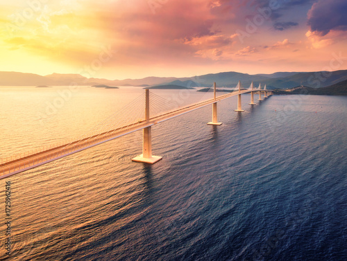 Aerial view of modern Peljesac Bridge and blue sea at colorful sunset in summer. Croatia. Top drone view of bridge during golden hour, mountains, reflection in water and orange sky. Architecture