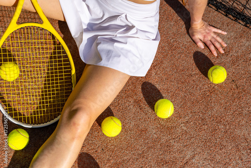 Female tennis player resting on the court with racket and scattered tennis balls in sunlight