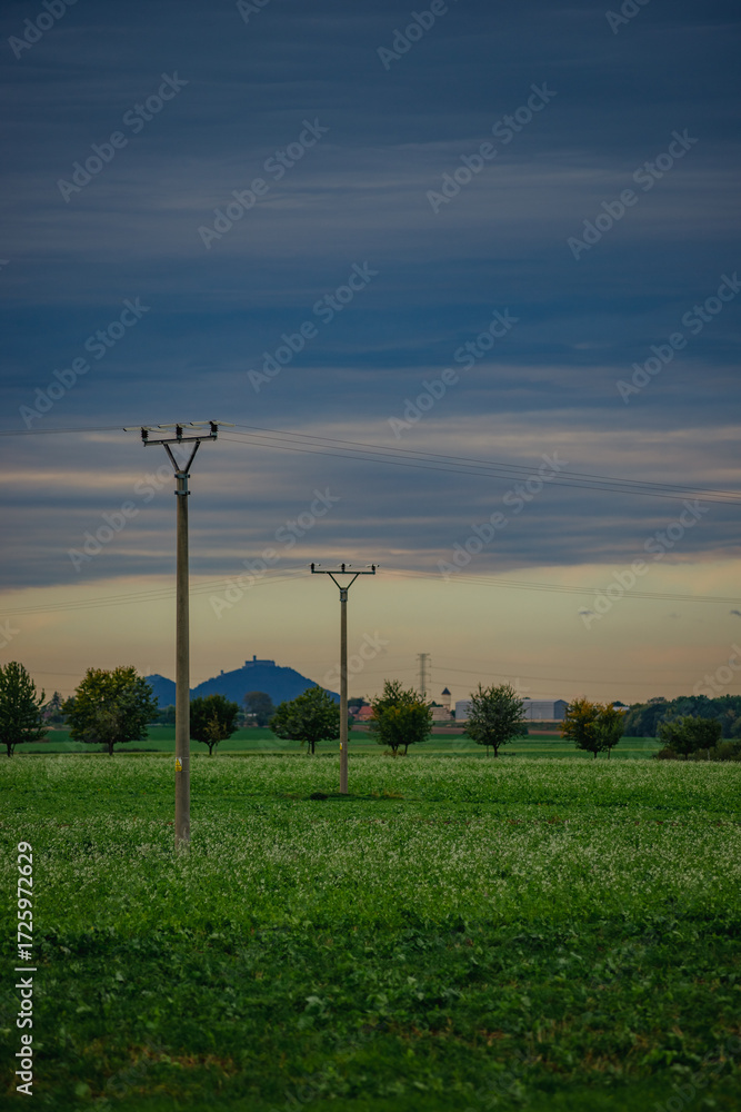 Obraz premium Minimalist rural landscape with power lines crossing a vast green field and a prominent hill with a fortress silhouette on the horizon under a soft blue and orange sunset sky.