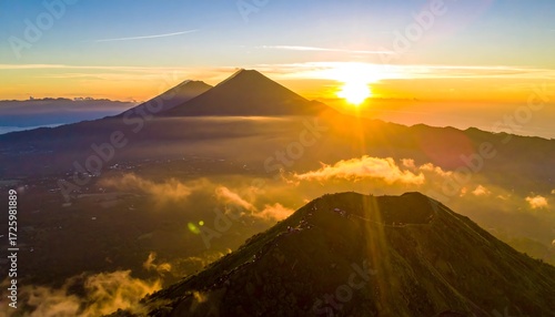 A breathtaking aerial view of three volcanic peaks bathed in the golden light of a rising sun, with scattered clouds below