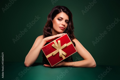 Amazing brunette woman holding a red gift box with gold ribbon against green background, celebrating holiday