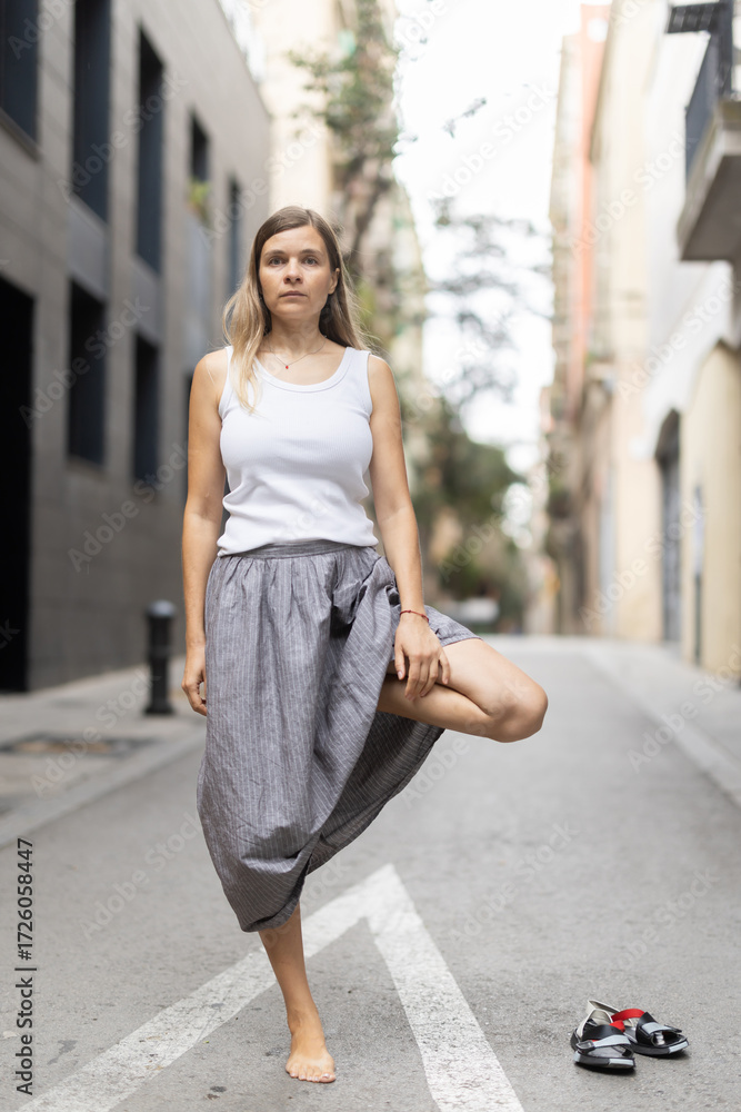 Fototapeta premium Female citizen in casual clothes perform tree yoga asana. Woman chose deserted urban highway for yoga break