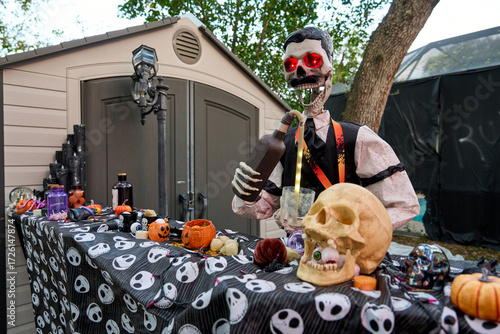 Animated skeleton with bright glowing eyes mixes a drink at a Halloween bar. Table filled with skulls, pumpkins, jars, and party decor in a backyard scene.