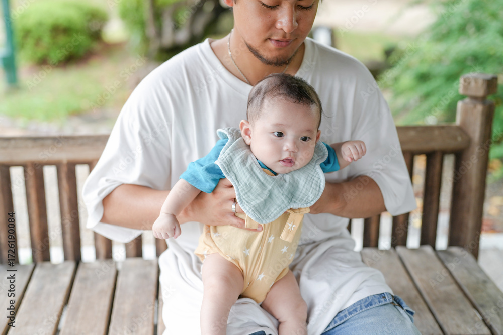 Fototapeta premium A man is holding a baby on a bench. The baby is wearing a blue shirt and a yellow outfit. The scene is peaceful and calm, with the man