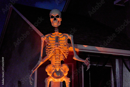 A towering skeleton figure stands by a house porch under orange spotlight. Spooky Halloween yard prop with bones and skull dominates the dark scene.