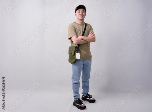 A young man stands with arms crossed, wearing a beige t-shirt, blue jeans, cap, and an olive crossbody bag in studio background