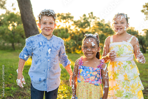 3 niños felices y sonrientes amigos hermanos bañados en espuma artificial al aire libre en un parque publico en un dia soleado disfrutando alegres de su inocencia
