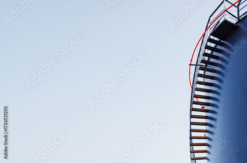 A close-up of dark blue industrial tank with a red safety rail on top, framed against a clear blue sky