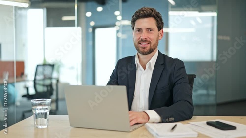 Professional businessman focused on laptop work in a modern office, wearing a sharp suit. Looks up, then smiles confidently, exuding success.