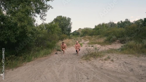 Two athletic men perform lunges on a sandy path in a lush natural setting, embracing an outdoor fitness routine. Healthy lifestyle and strength training in nature.