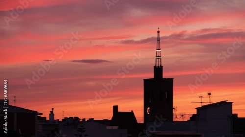 Silhouette of a city skyline at sunset. The sky is filled with vibrant orange and pink hues. A tall tower stands prominently against the colorful backdrop.