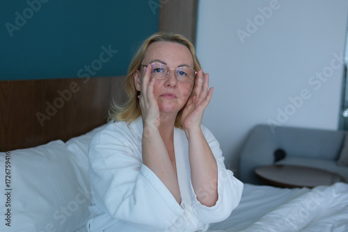woman applying sheet mask at home in front of double sided mirror.beautiful girl in white robe sitting with coffee or tea cup in hand. relax and wellbeing moment. lot of cosmetic in front of female.