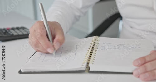 Close-up of hands - a man in a shirt draws a diagram and takes notes with a pen in a notepad on a metal spring on an office desk