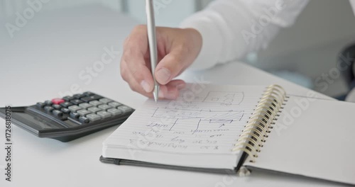 Close-up of hands - a man in a shirt draws a diagram and takes notes with a pen in a notepad on a metal spring on an office desk