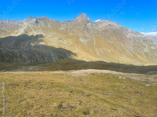 Expansive mountain landscape in the Mont Avic Natural Park, located in the Aosta Valley, Italy. The foreground features a grassy plateau with patches of bare earth, while dramatic rocky peaks rise