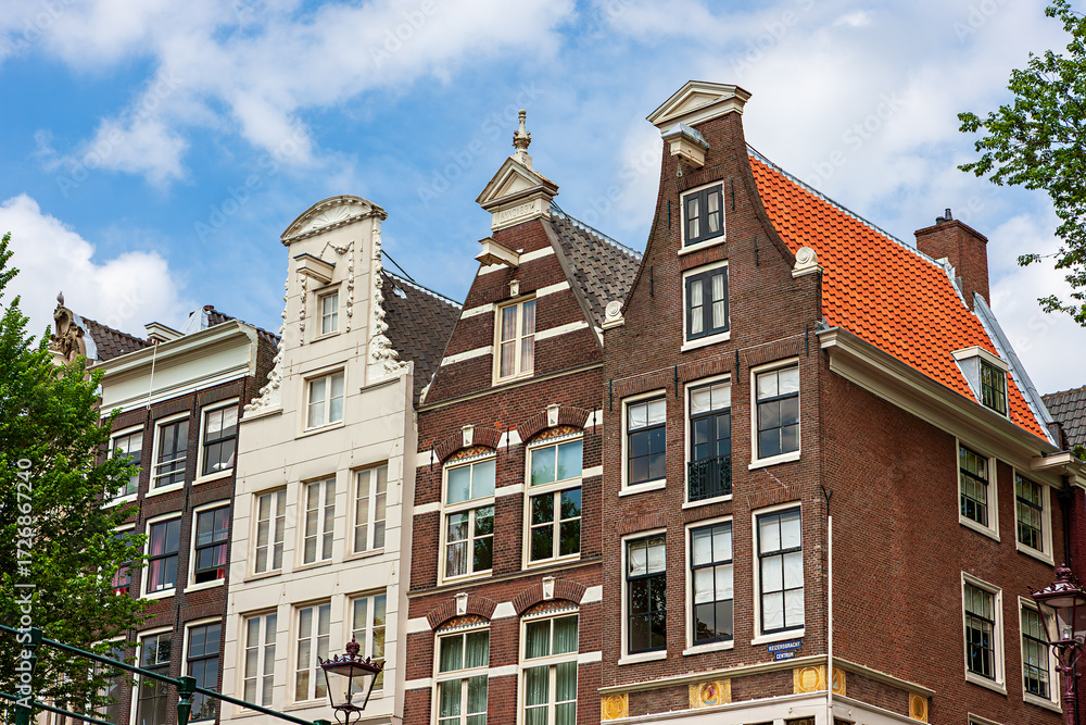 Fototapeta premium In Amsterdam, the charming facades of colorful houses showcase classic Holland architecture. The buildings feature unique designs and vibrant colors against a bright blue sky.