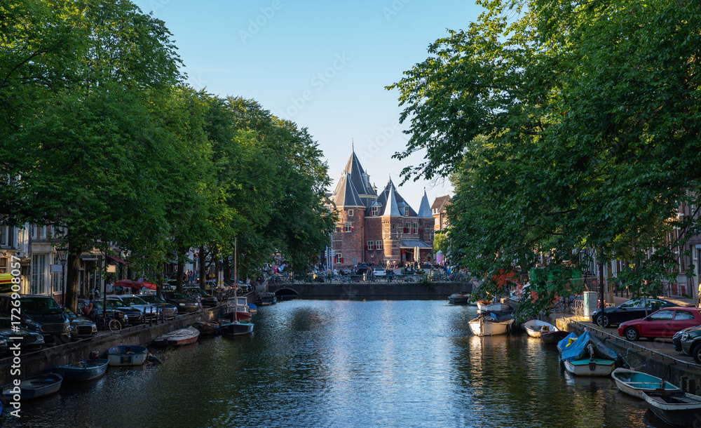 Naklejka premium The Waag, a medieval city gate turned museum and restaurant, stands at the end of a canal in Amsterdam, Netherlands, framed by green trees, moored boats, and parked cars on a summer day.