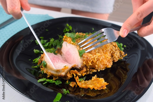 Woman hands cutting breaded chicken breast cutlet serving with greenery on black plate. Tasting cooked golden-brown crispy chicken poultry fillet. Culinary cuisine recipe, prepare food dish.