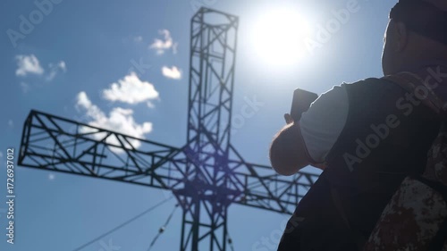 Rear view of an overweight man sitting on a mountain peak near a tall metal cross, filming the Christian cross with a smartphone held in both hands. A moment of solitude and prayer to God.