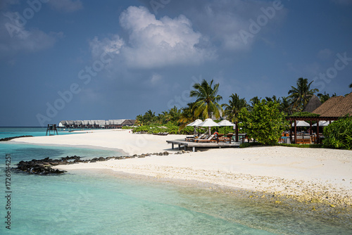 Fototapeta Naklejka Na Ścianę i Meble -  White sandy beach on a Maldivian island with turquoise water, over water villas in the distance, palm trees, sun loungers with umbrellas, and volcanic rocks under a dramatic tropical sky.