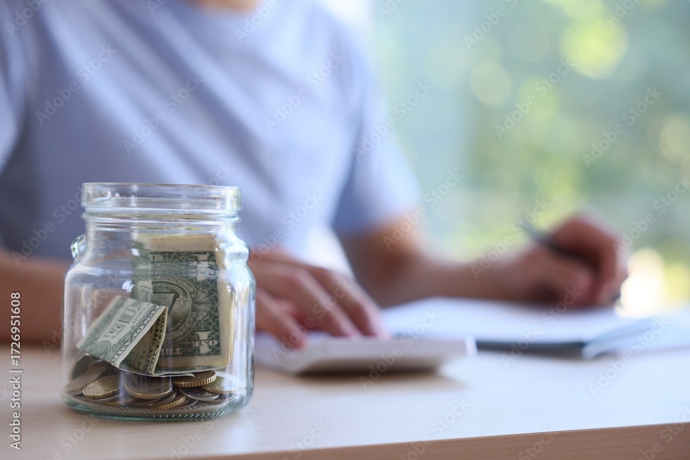 Fototapeta premium Glass jar with money and man budgeting at wooden desk, selective focus