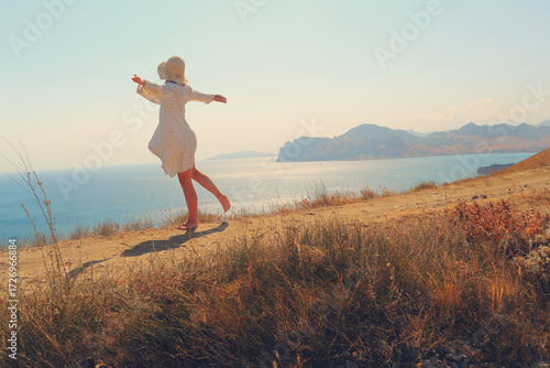 A girl in a dress and a hat stands in the sun, blown by the wind, on the seashore in the heat