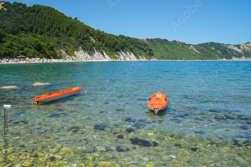 Sunny day at the beach across the cliffs in Portonovo Beach, Marche, Italy. Two red boats on the waves across the cliffs and sea
