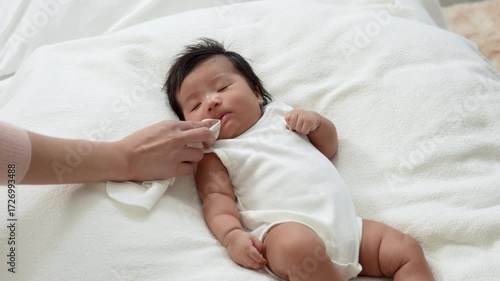 Asian newborn baby girls wearing a white bodysuit laying on the bed, mother hand a napkin clean her mouth.
